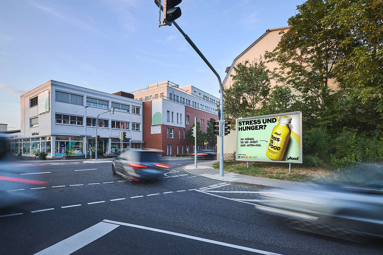 Großes yfood-Werbeplakat mit der Aufschrift „Stress und Hunger?“ an einer städtischen Kreuzung mit vorbeifahrenden Autos und Gebäuden im Hintergrund.