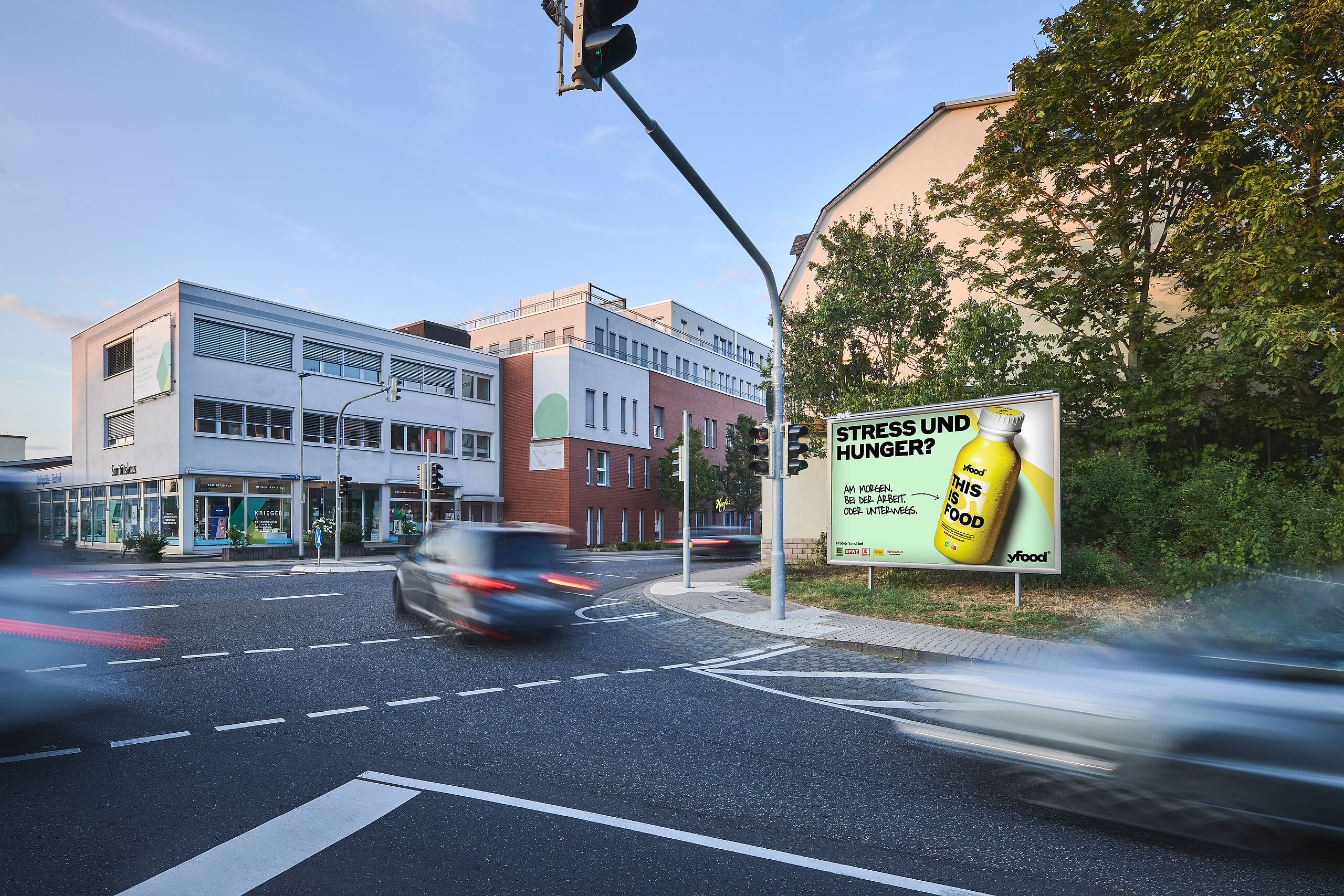 Großplakat an stark befahrener Kreuzung in der Stadt Großes yfood-Werbeplakat mit der Aufschrift „Stress und Hunger?“ an einer städtischen Kreuzung mit vorbeifahrenden Autos und Gebäuden im Hintergrund.