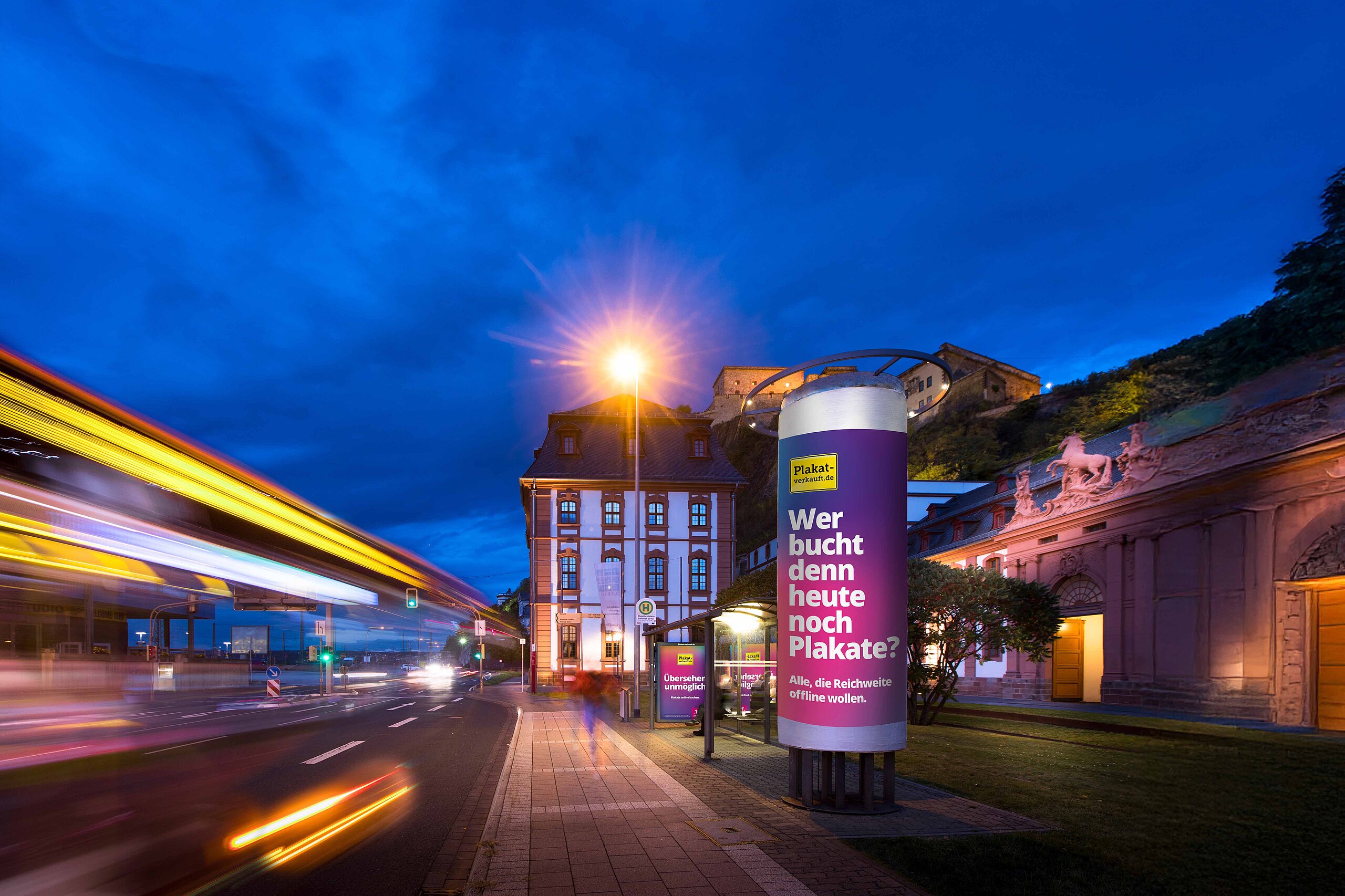 Ganzsäule mit Werbebotschaft von Plakat-verkauft.de bei Nacht, beleuchtet mit Verkehr auf der Straße und historischem Gebäude im Hintergrund.