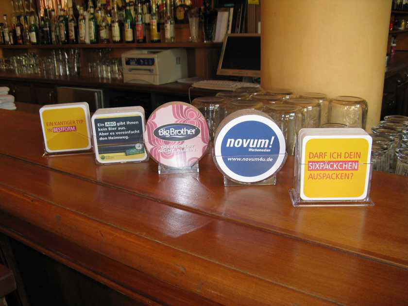 Mehrere bedruckte Bierdeckel in Aufstellern auf einer Bar-Theke, genutzt als Werbeflächen für verschiedene Kampagnen in der Gastronomie.