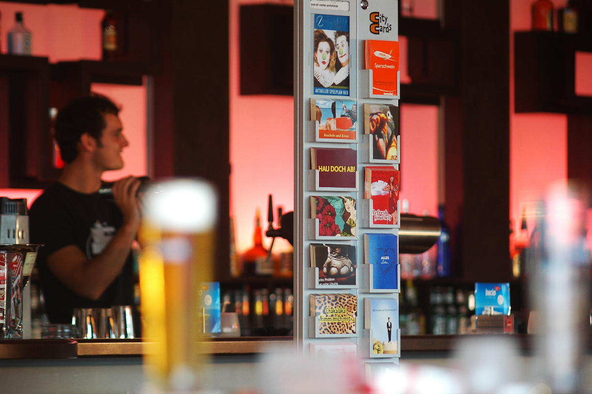 Postkartenständer mit bunten Werbepostkarten in einer Bar, im Hintergrund ein Barkeeper und die beleuchtete Tresenlandschaft.